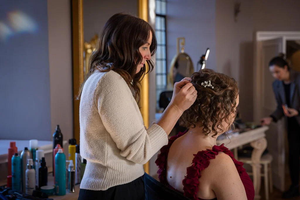 A bridal stylist meticulously places small white flower pins into the curly brown hair of a bride wearing a maroon ruffled dress during morning preparations.
