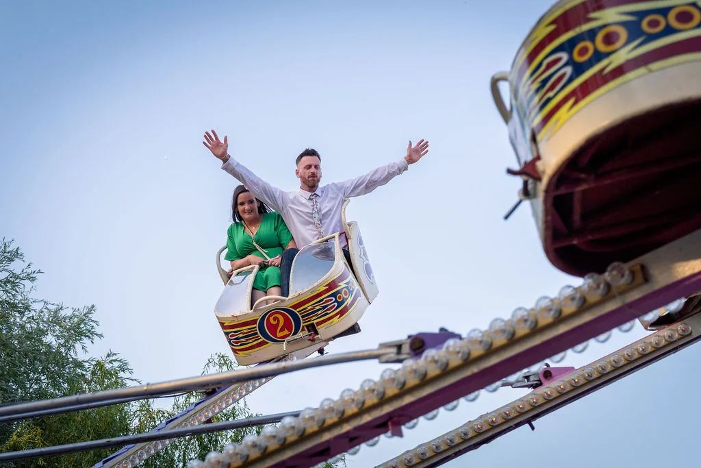 A newlywed couple excitedly ride a vintage fairground attraction at their festival-themed wedding, with the groom raising his arms in the air against a clear blue sky.