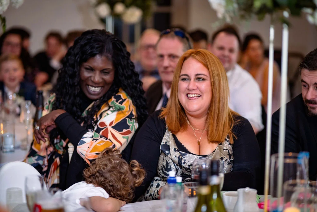 Two smiling guests laugh joyfully during wedding speeches at a reception table while a young child rests nearby.