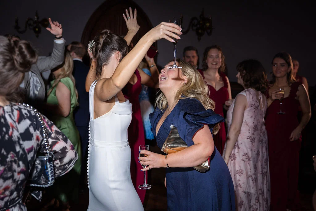 A laughing bride in a sleek white gown dances joyfully with wedding guests holding champagne glasses during the evening reception.