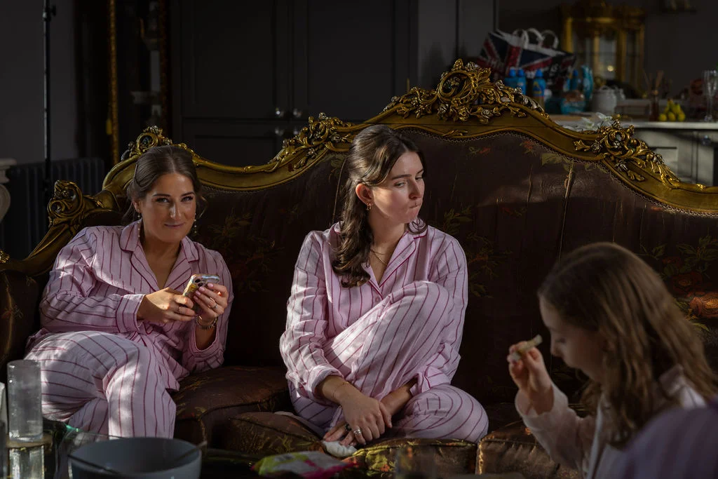 Two bridesmaids relax on an ornate gilt sofa in matching pink striped pyjamas during bridal morning preparations.