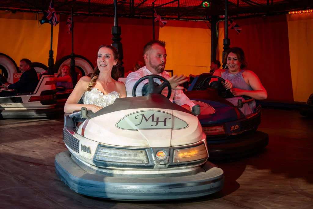 A bride in a strapless dress and a groom laughing as they drive a bumper car together at a funfair themed evening wedding reception.