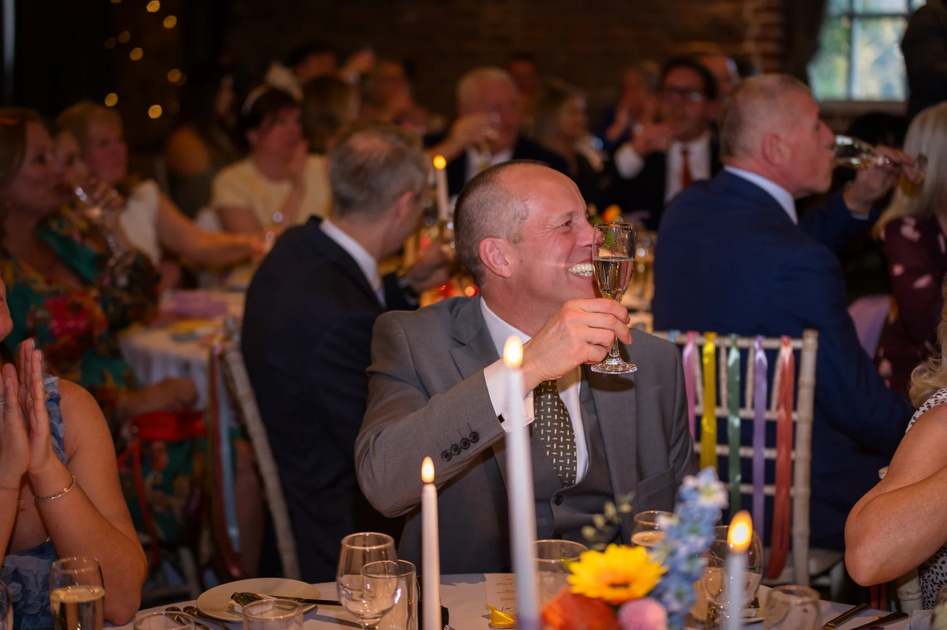 A wedding guest in a grey suit raises a glass of champagne with a joyful laugh during the reception speeches.