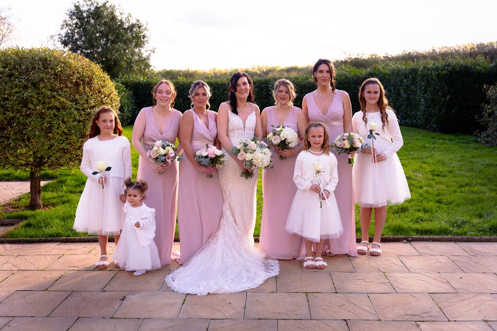 A smiling bride in a lace wedding gown poses outdoors on a stone patio with her adult bridesmaids in pink dresses and young flower girls holding single roses.