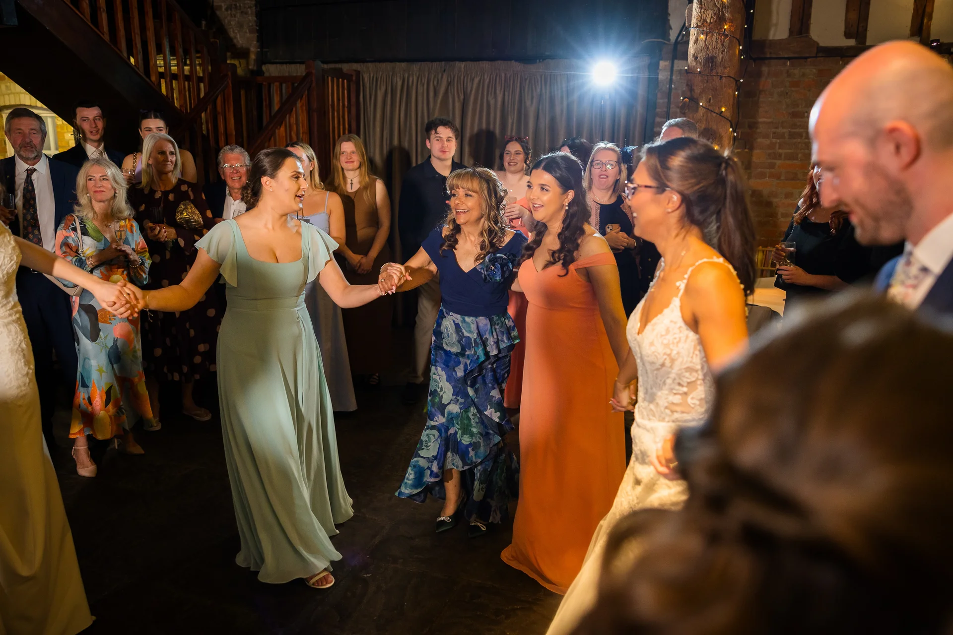 Wedding guests join hands in a lively dance circle during the evening reception inside a rustic barn with exposed brick walls.