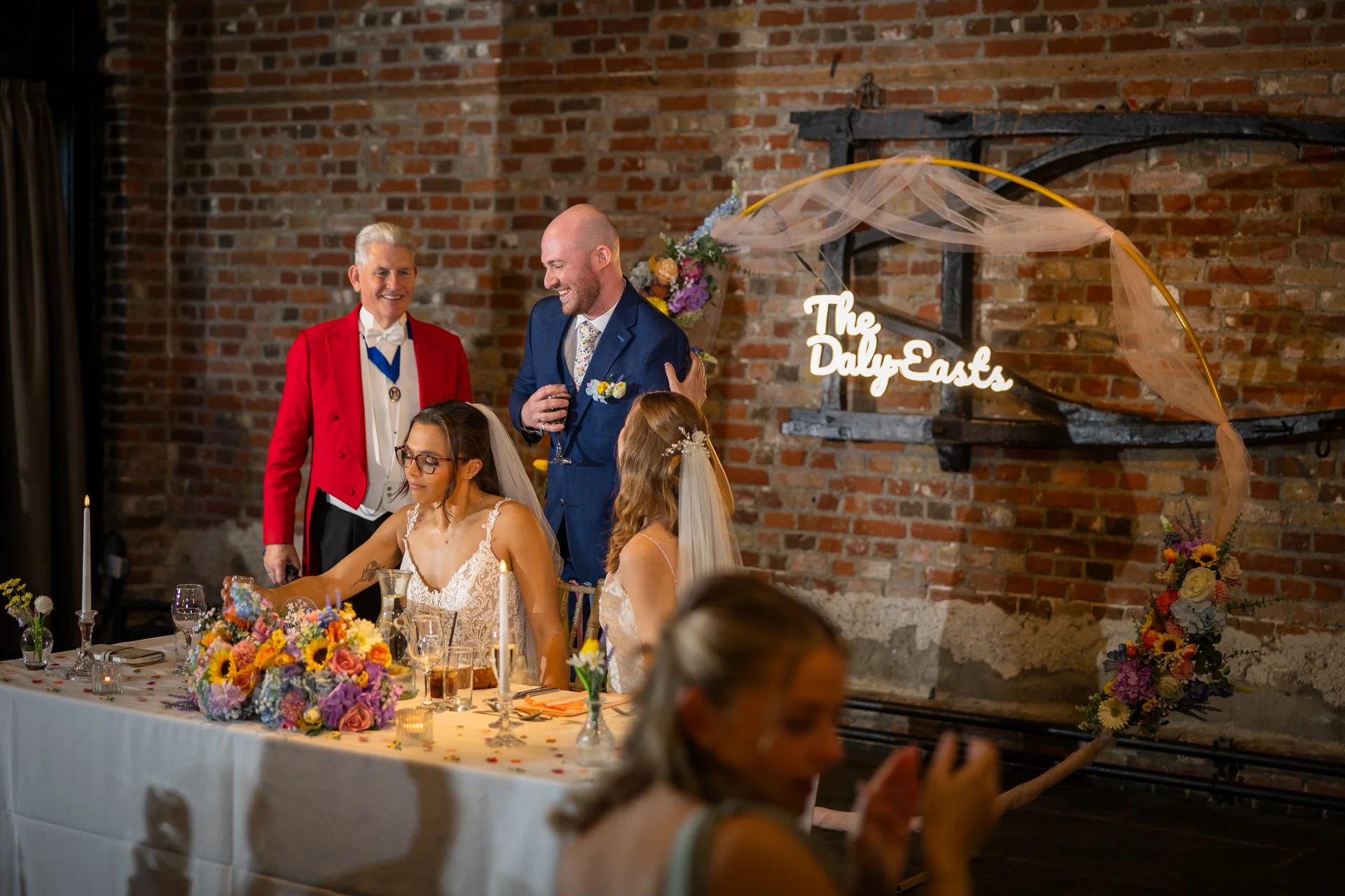 A smiling couple and guests share a toast during wedding speeches at a reception table decorated with colourful florals and candles against an exposed brick wall.