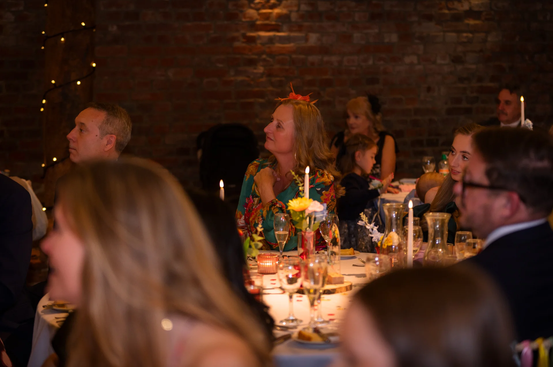 Wedding guests gathered around a candlelit dining table with exposed brick in the background, listening attentively during the wedding reception speeches.