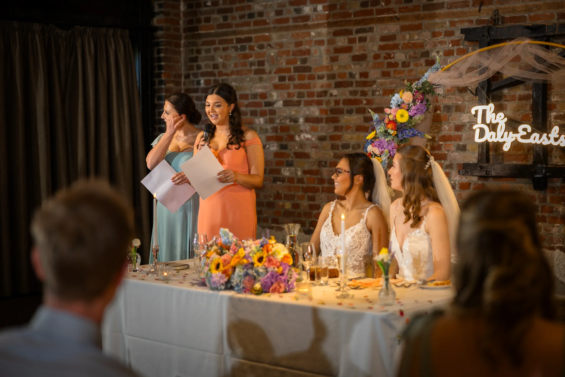 A smiling bridesmaid delivers a heartfelt speech during the wedding reception while two brides and another bridesmaid listen from the top table in a rustic brick venue.
