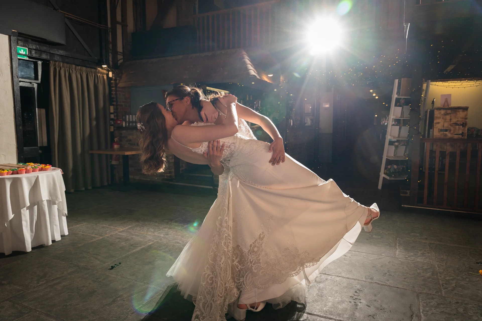 Two brides share a dramatic dip and kiss during their first dance inside a rustic barn illuminated by bright backlighting and overhead string lights.