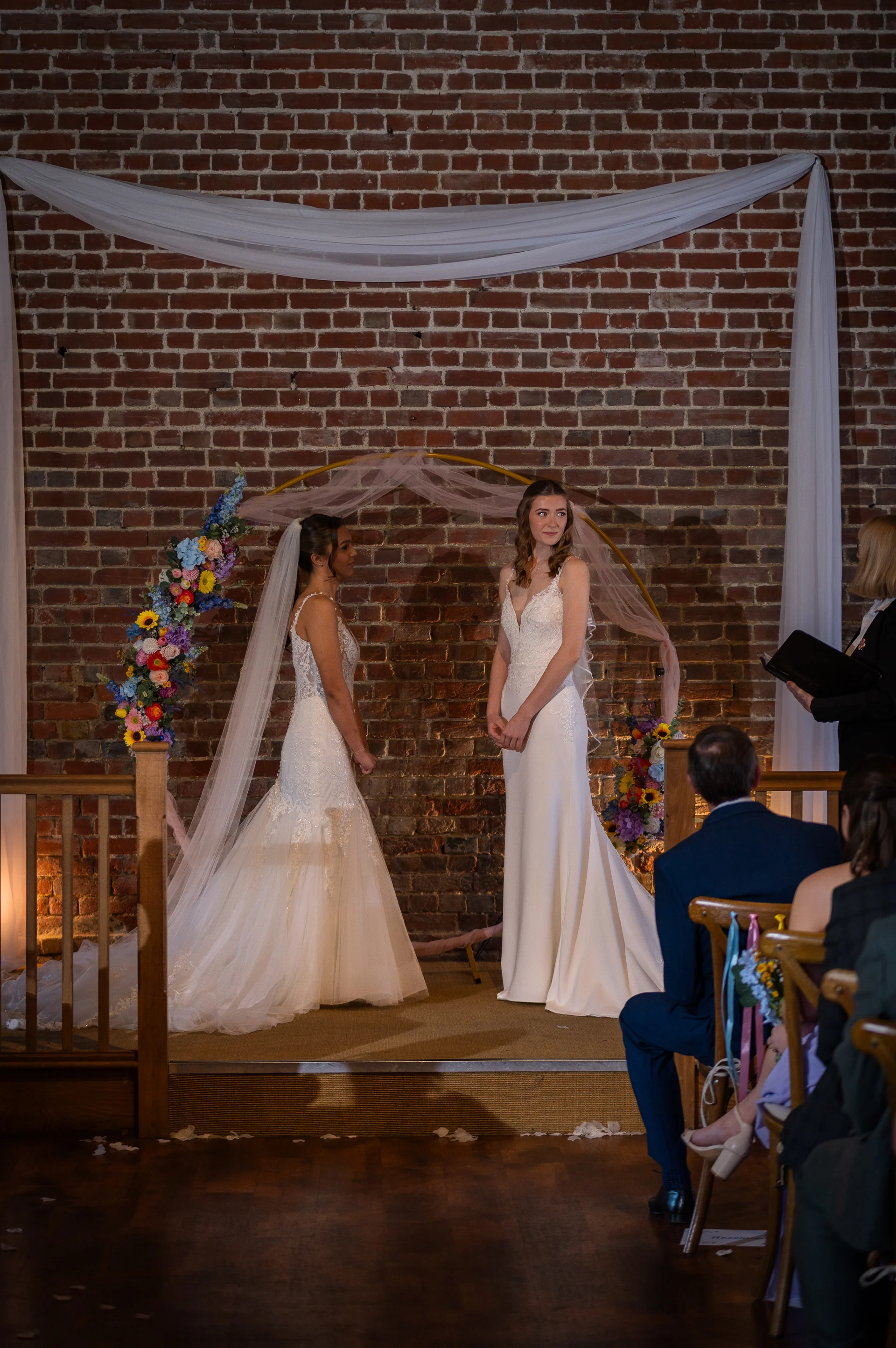 Two brides stand hand in hand during their wedding ceremony in front of an exposed brick wall and a floral arch.