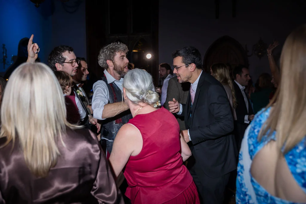 A candid photograph capturing wedding guests laughing and dancing together during an evening reception illuminated by direct flash.