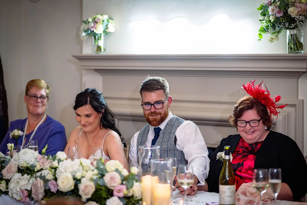 The newly married couple and family members seated at the top table during wedding speeches, with floral arrangements and candlelight adorning the mantelpiece.