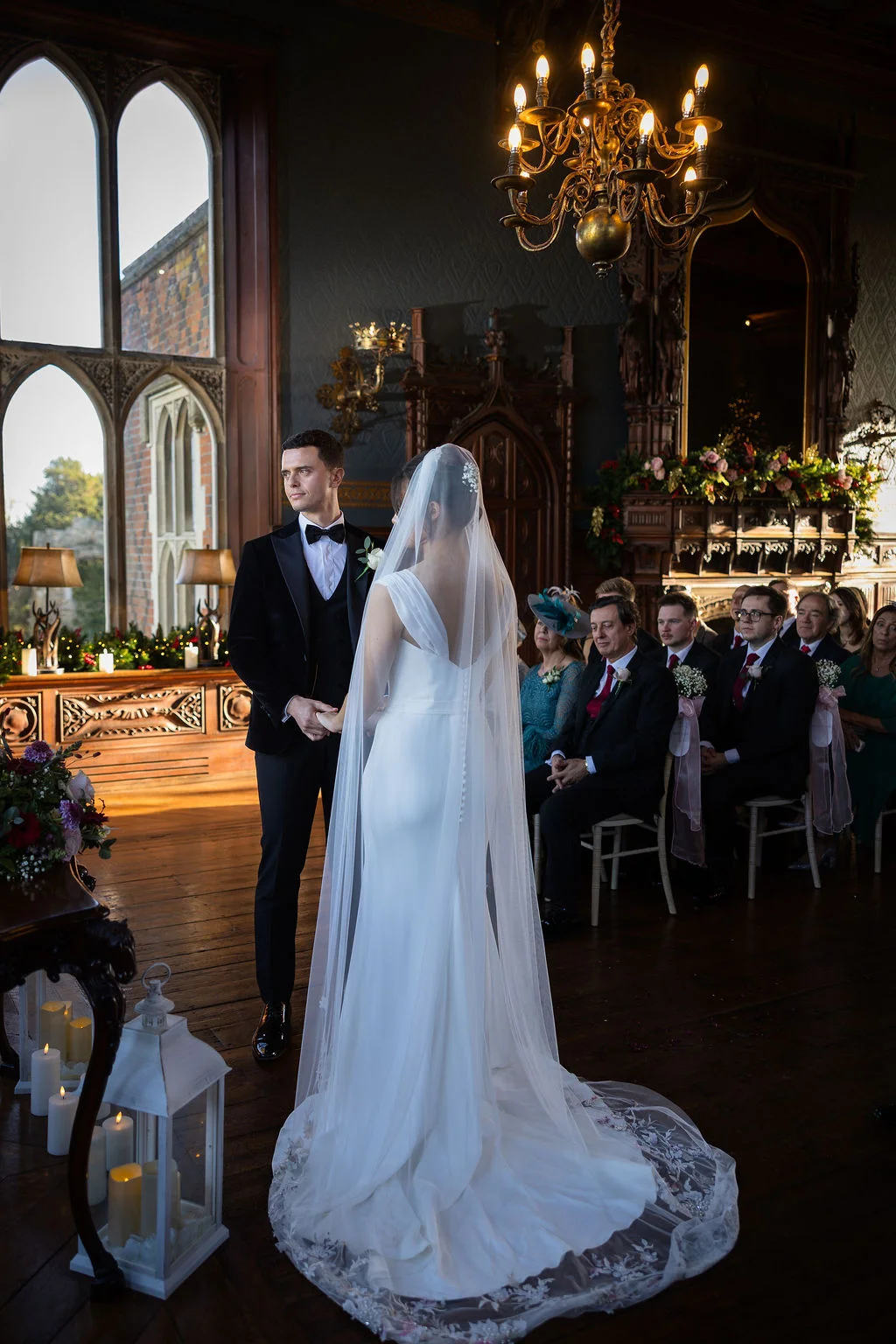 A bride in a sleek crepe gown and embroidered chapel veil stands facing her groom in a black tuxedo during an indoor wedding ceremony within a grand panelled hall.