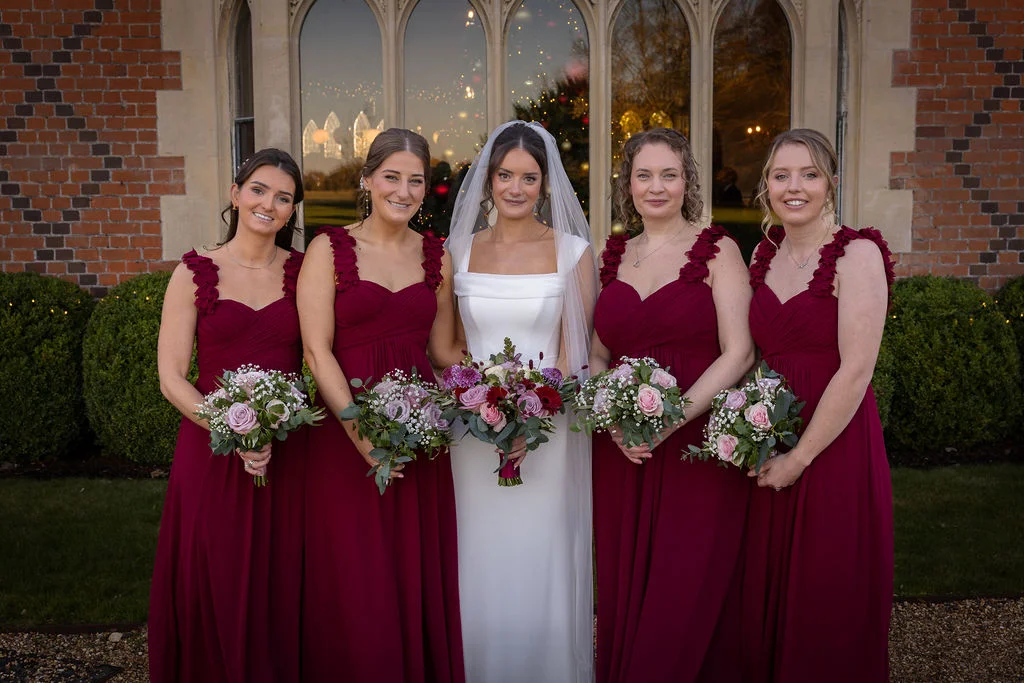 A beautiful bride in a sleek white gown and veil stands proudly with her four bridesmaids in matching burgundy dresses, holding bouquets outside a historic brick building.