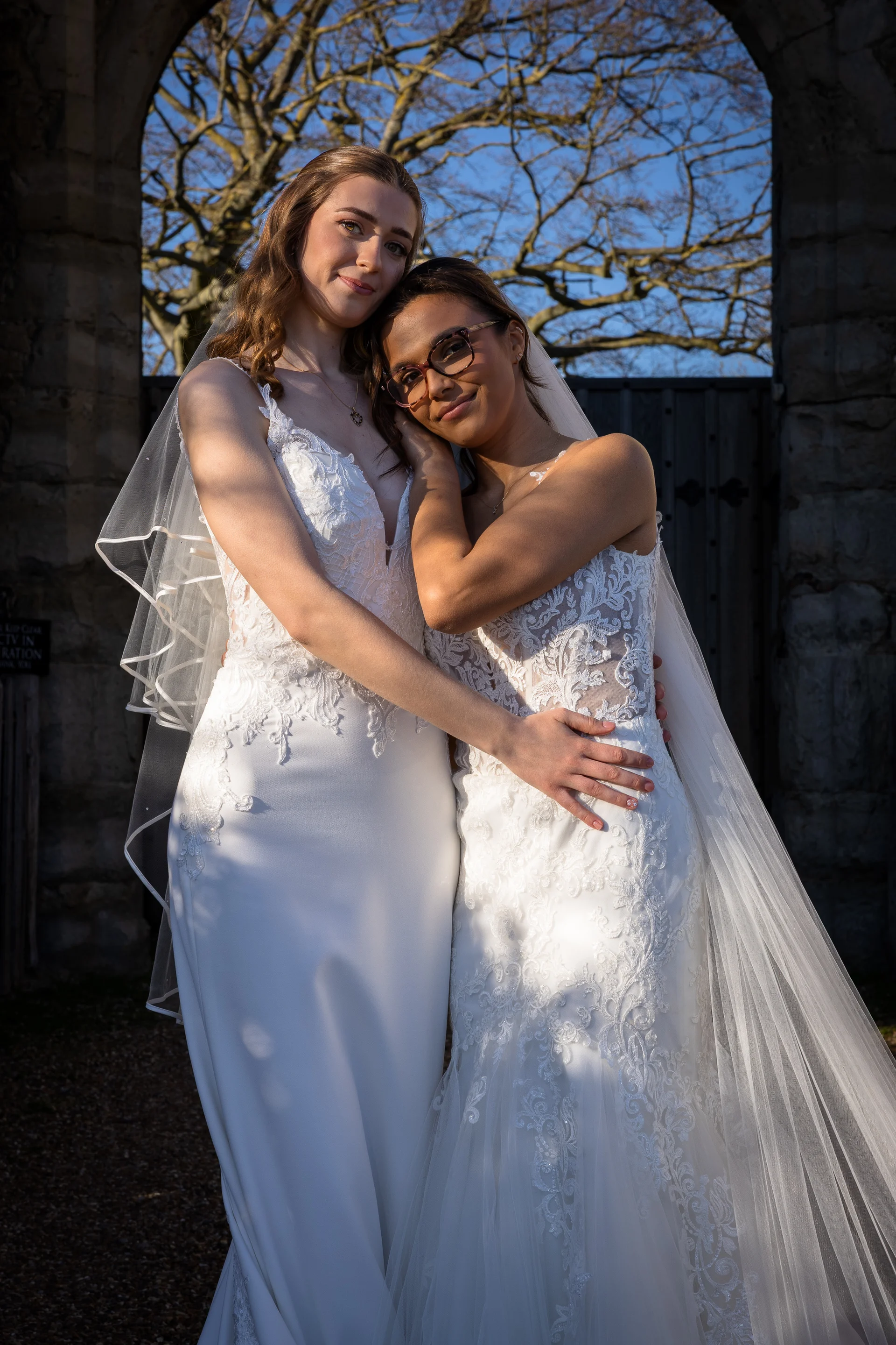 Two smiling brides in gorgeous lace wedding gowns embrace warmly within a historic stone archway during golden hour.