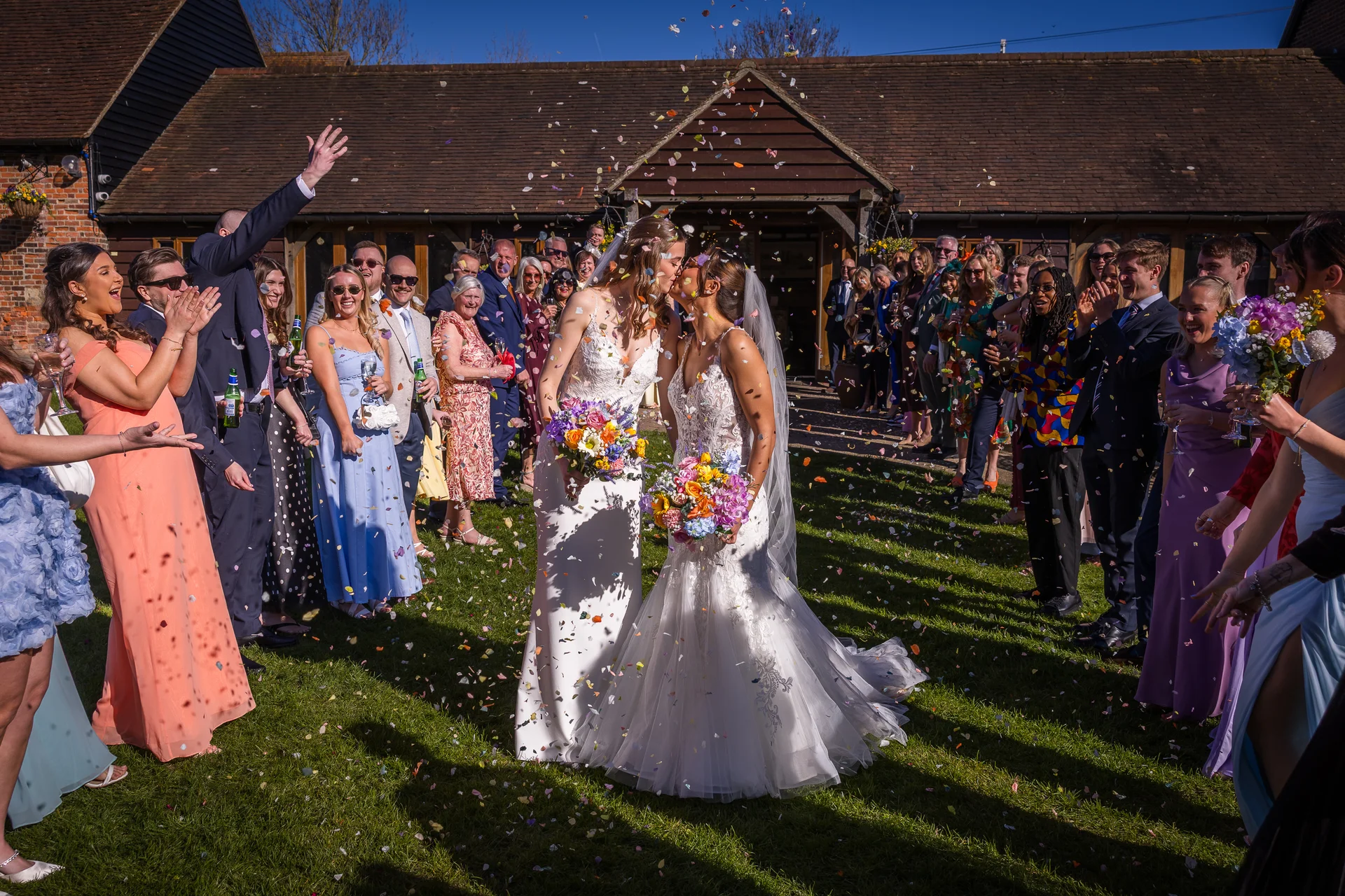 Two brides share a romantic kiss on a sunny lawn outside a rustic barn as wedding guests shower them with colourful confetti.