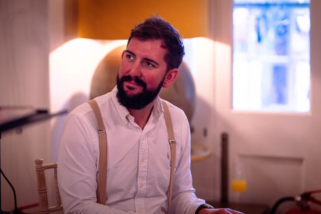 A bearded groom sits indoors wearing a white shirt and braces, looking off to the side with soft window light illuminating the scene.