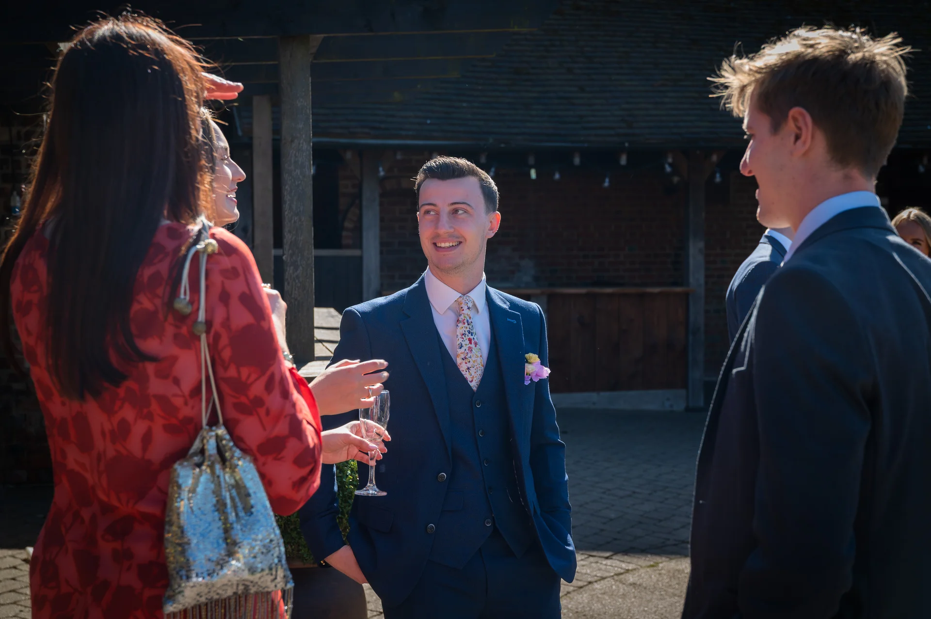 Wedding guests mingling outdoors in bright sunlight during a drinks reception, with a smiling groom in a navy blue three-piece suit holding a glass of champagne.