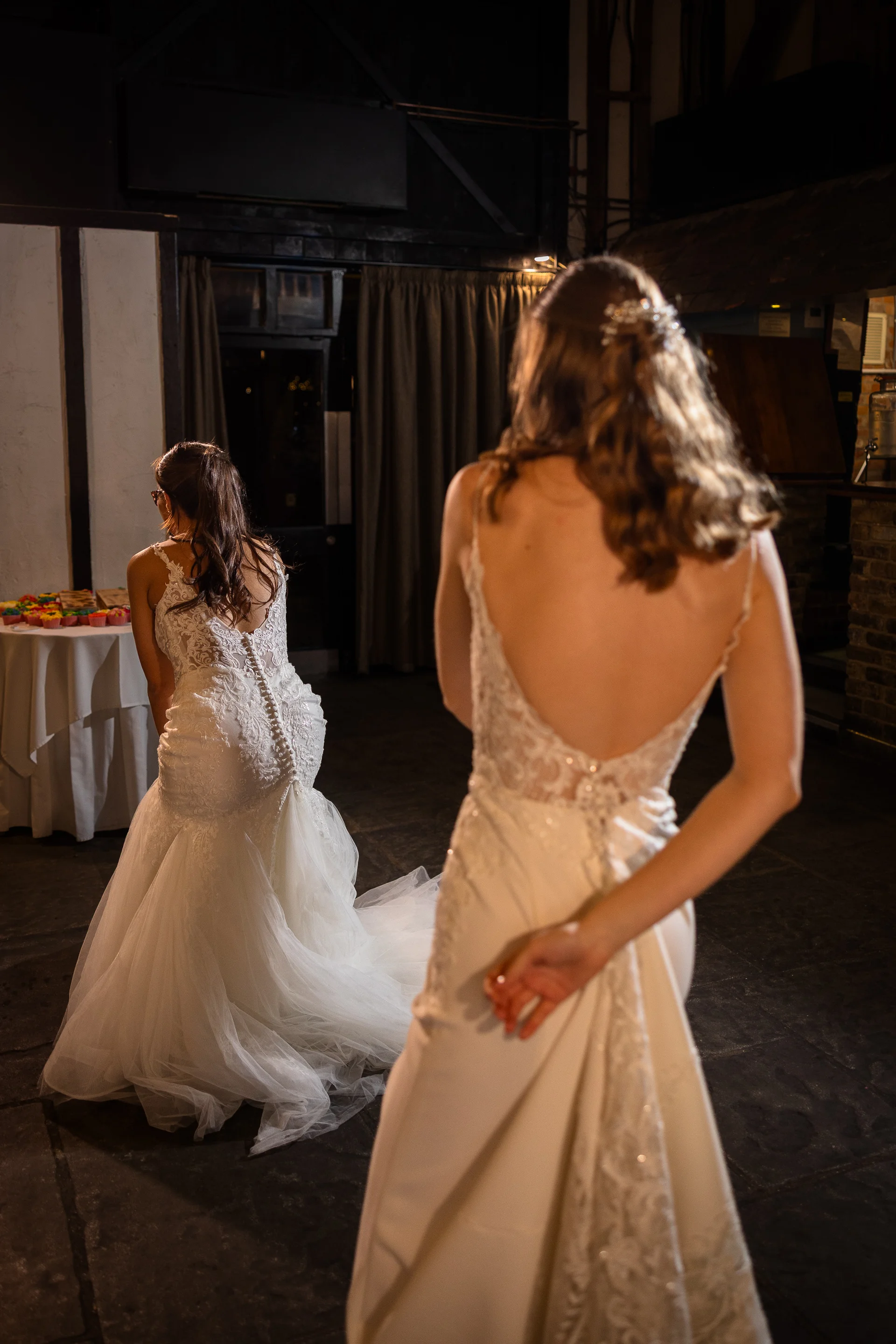 Two brides stand back to back indoors wearing detailed lace and tulle wedding dresses with low backs and flowing trains.