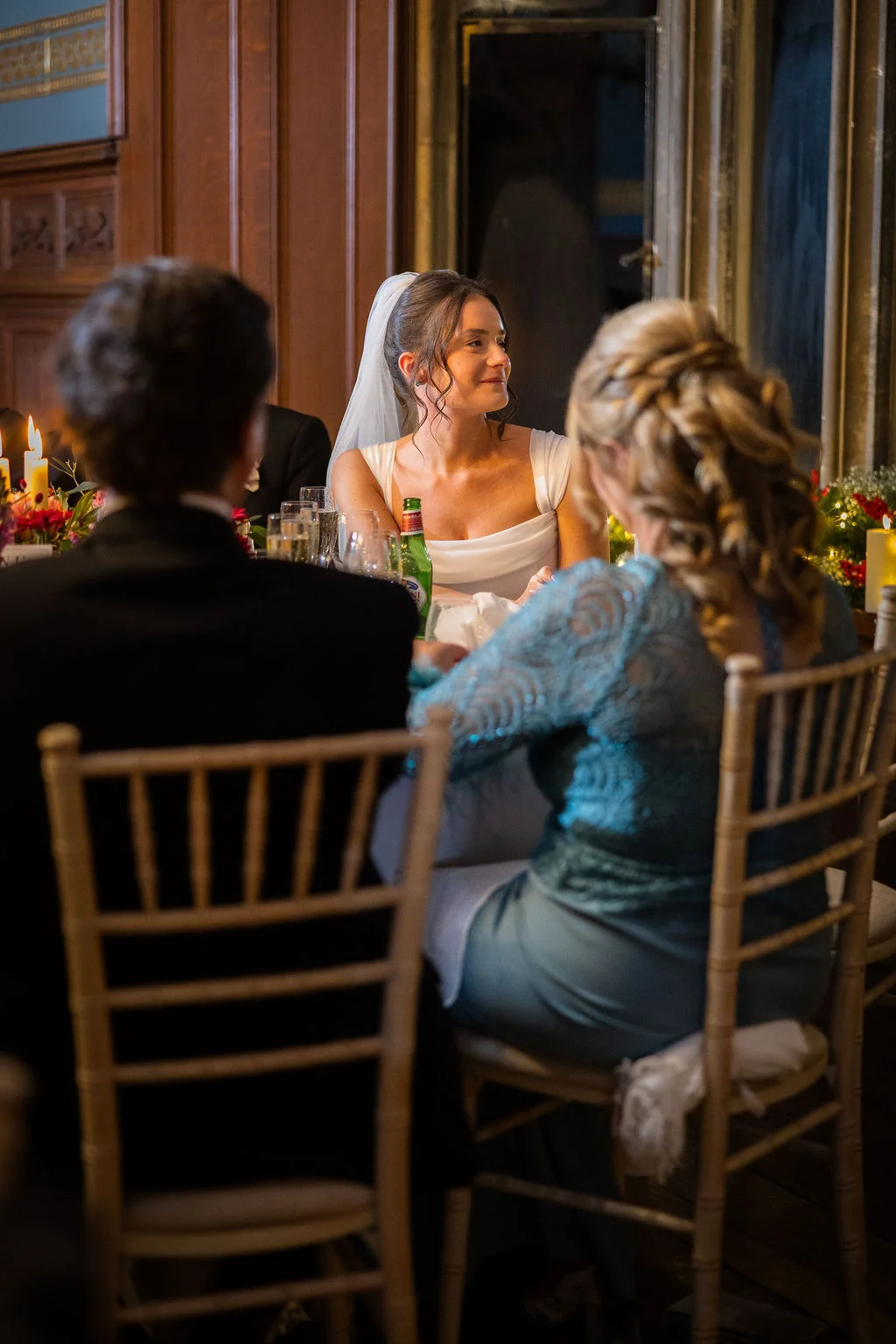 A smiling bride sits at the top table during the wedding reception in a warm, panelled room illuminated by candlelight.