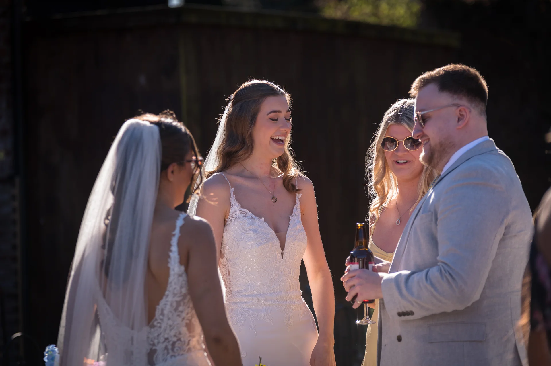 A bride in a lace wedding dress shares a joyful laugh with friends outdoors in bright sunlight during the wedding reception.