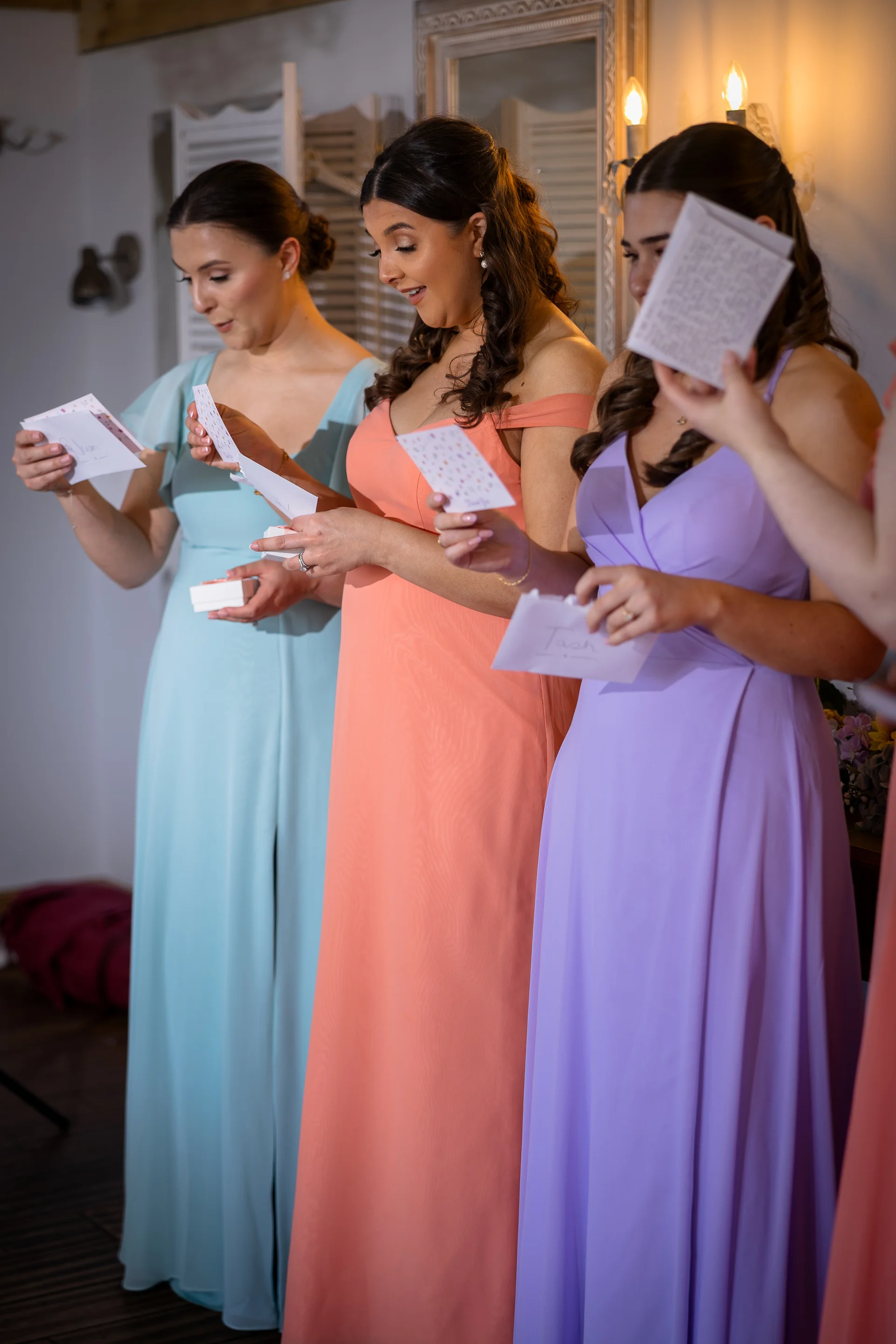 Three smiling bridesmaids in colourful pastel dresses read heartfelt letters together during morning bridal preparations indoors.