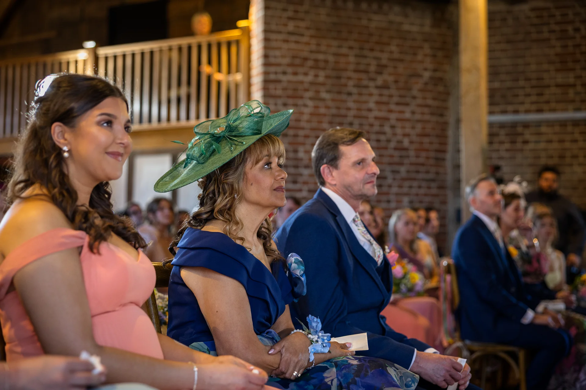 Wedding guests seated inside a rustic barn with exposed brick walls, watching the ceremony attentively.