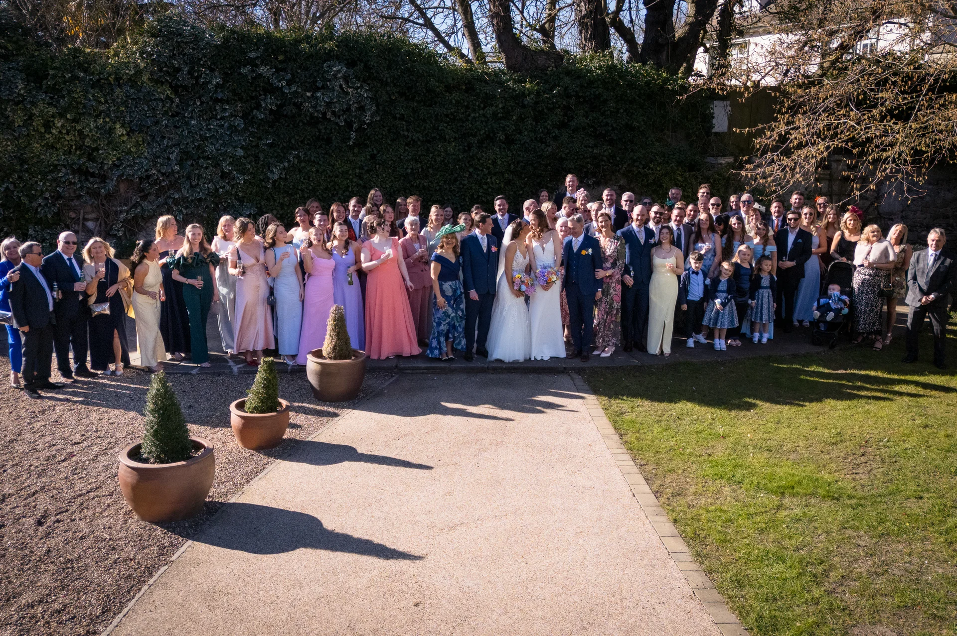 A large wedding party and guests gather for a group photograph outdoors in a manicured garden lined with potted plants and an ivy-covered stone wall.