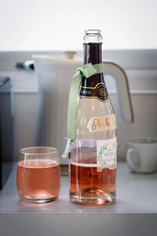 A close-up shot of a bottle of pink sparkling wine decorated with a pale green ribbon, sitting next to a glass of the same pink drink on a white surface.