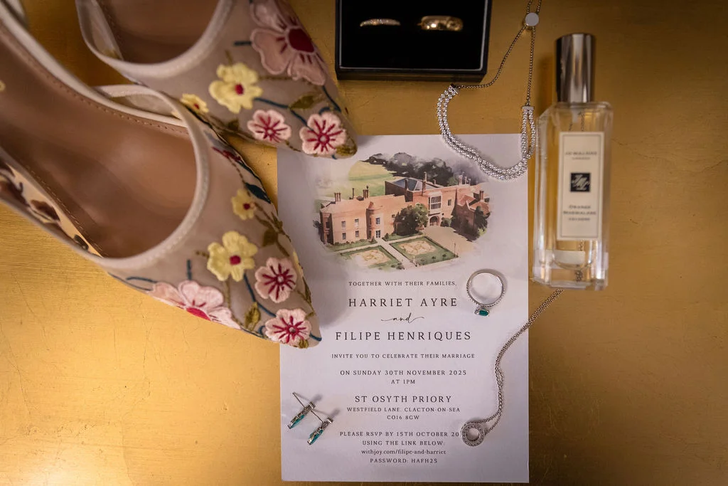 An elegant bridal flat lay showcases an illustrated wedding invitation suite for St Osyth Priory alongside floral embroidered heels, wedding bands in a velvet box, delicate jewellery, and a bottle of perfume on a gold surface.