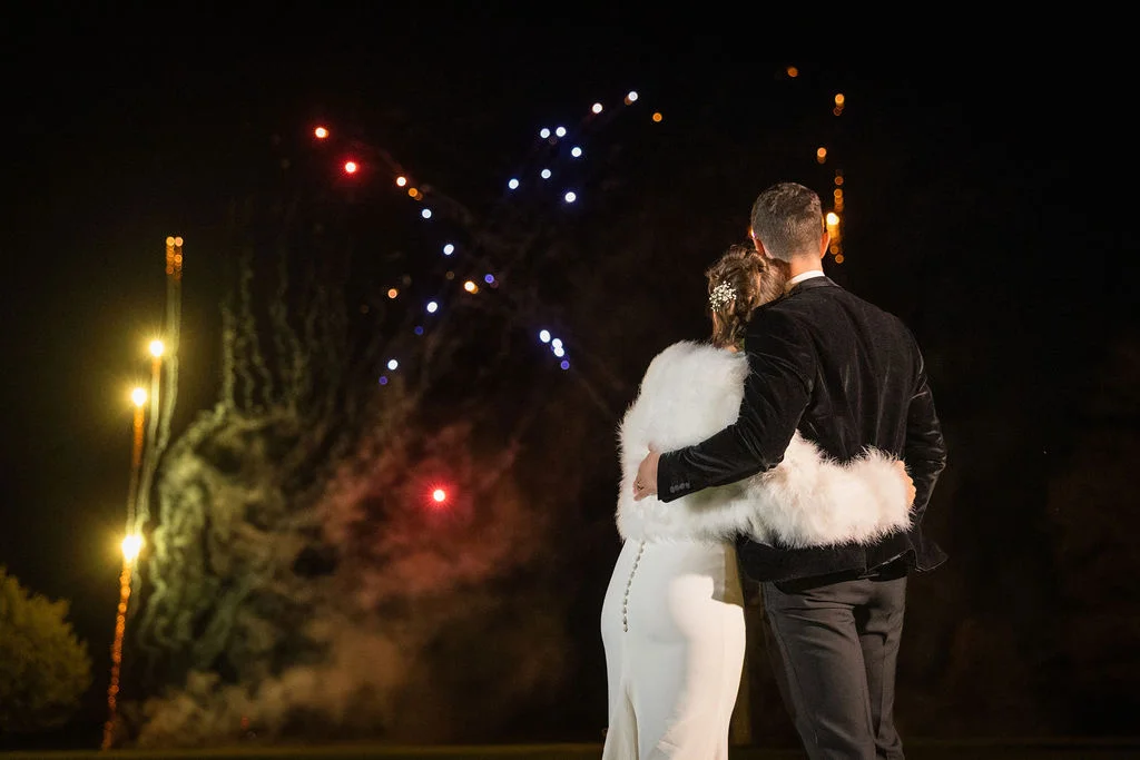 A newlywed couple embraces from behind while watching a vibrant fireworks display light up the night sky during their winter wedding celebration.