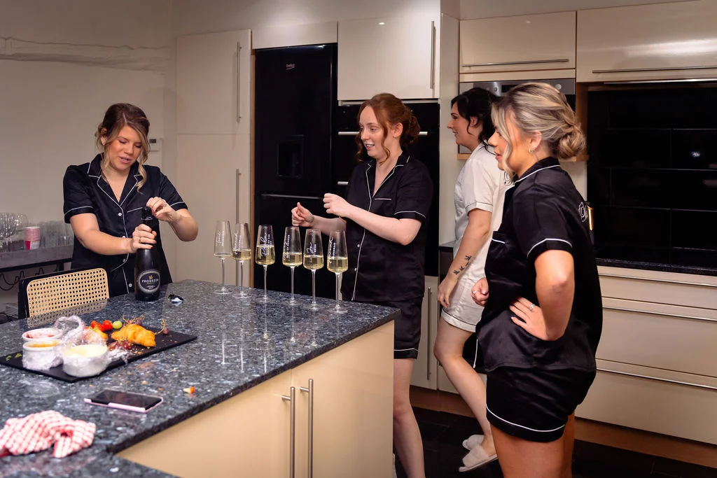 Four bridesmaids wearing matching black silk pyjamas gather around a kitchen island, laughing while opening a bottle of champagne and pouring drinks during morning bridal prep.