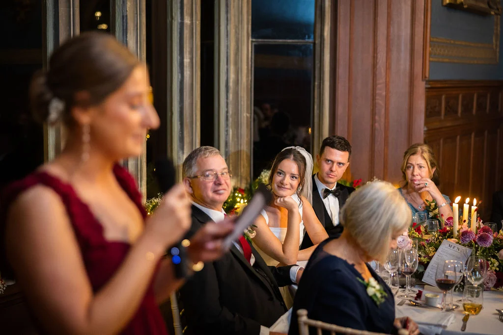 A bridesmaid delivers a speech at the top table while the bride, groom, and family members listen attentively in a candlelit, wood-panelled reception room.