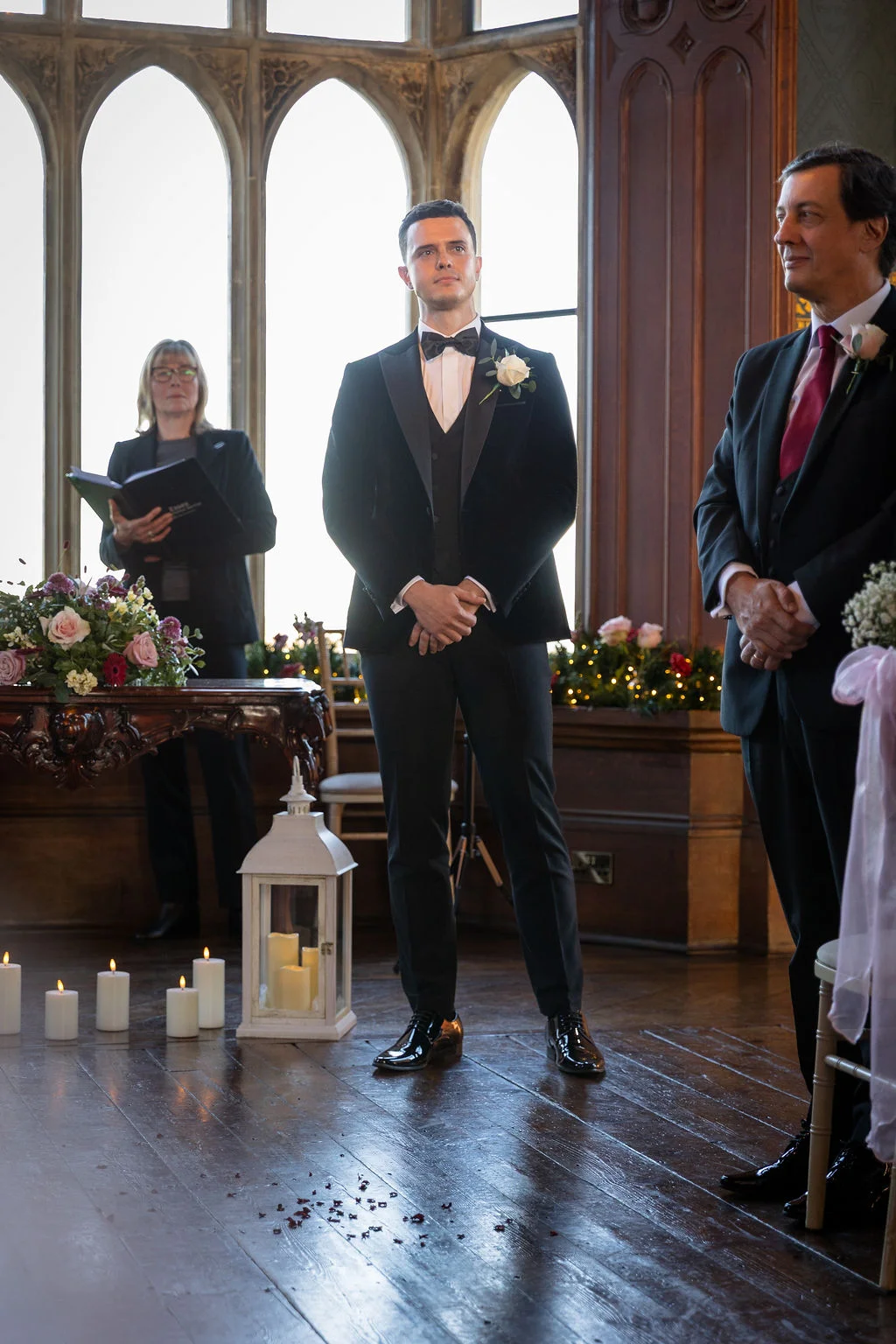 A handsome groom in a black velvet tuxedo waits at the altar during a candlelit civil ceremony inside a historic country house with large arched windows.