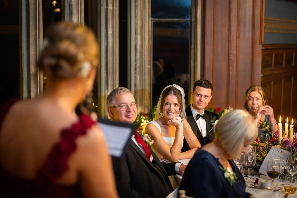 The smiling bride listens to a speech at the top table during an elegant evening wedding reception in a panelled hall.