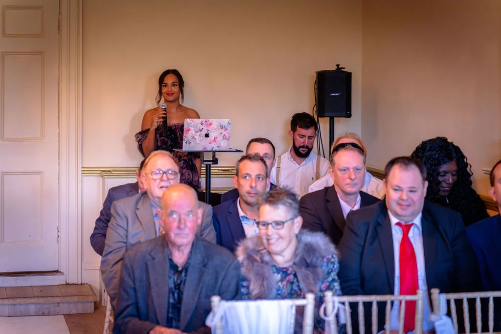 A woman stands and speaks into a microphone during wedding speeches while seated guests look on attentively.