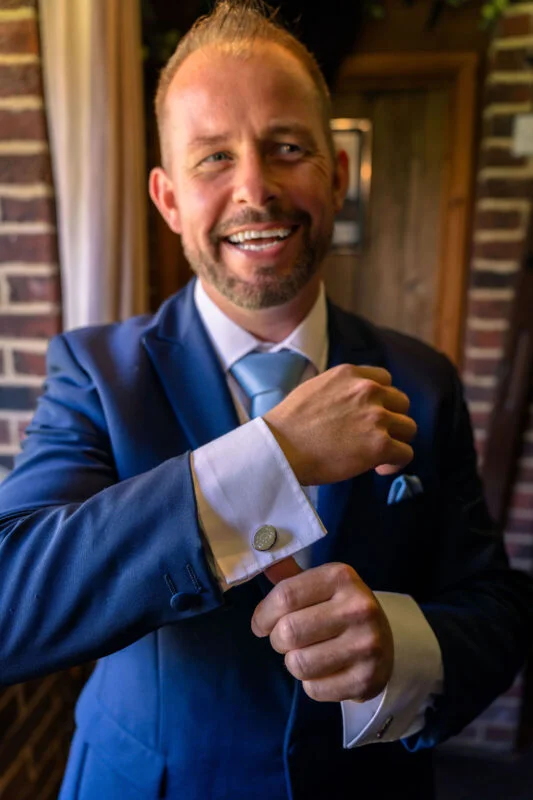 A smiling groom wearing a navy suit and light blue tie adjusts his cufflinks while getting ready near an exposed brick wall.