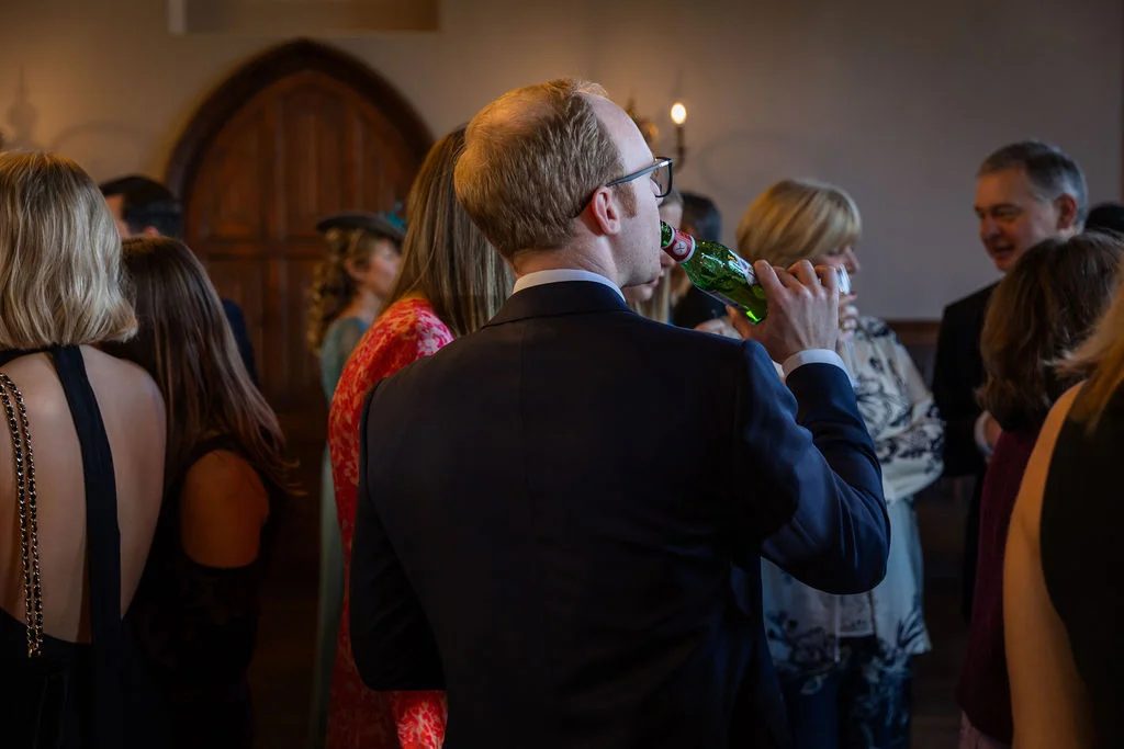 A guest in a dark suit drinks from a green glass bottle while mingling with others at a wedding reception.