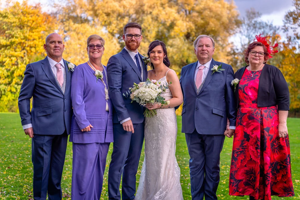 A bridal couple poses outdoors with their family on a green lawn surrounded by autumn trees with golden foliage.