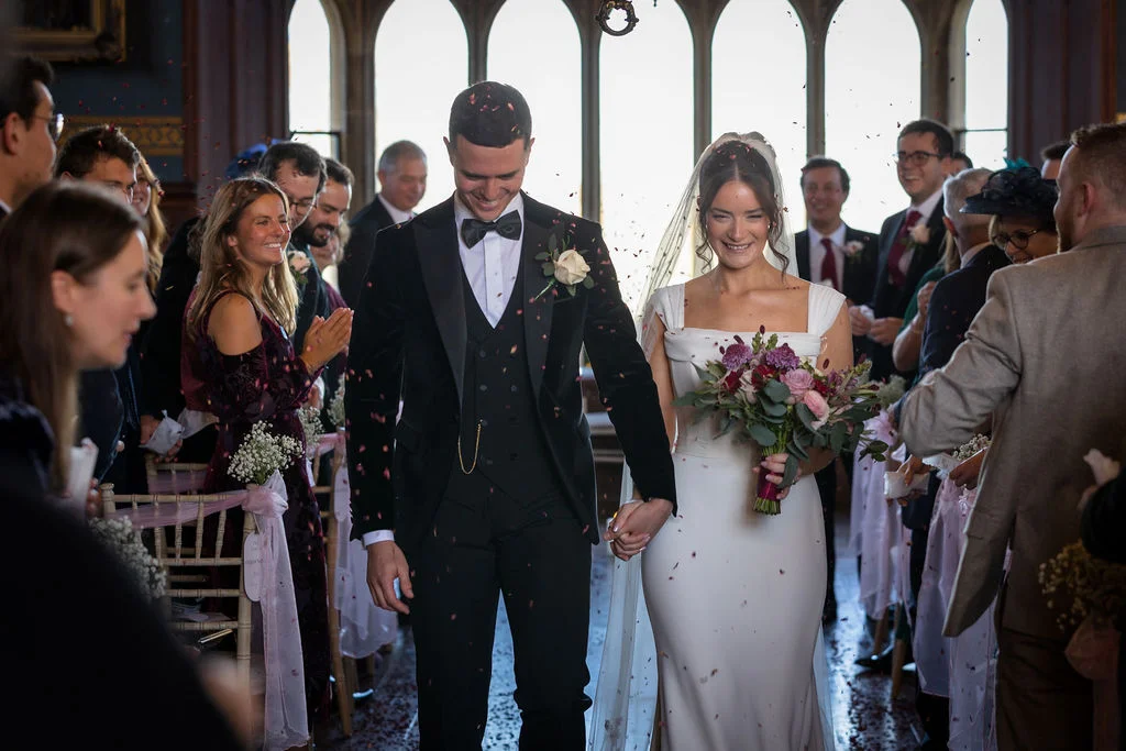 The newlywed couple joyfully walk up the aisle holding hands while guests shower them with confetti in a historic venue with tall windows.