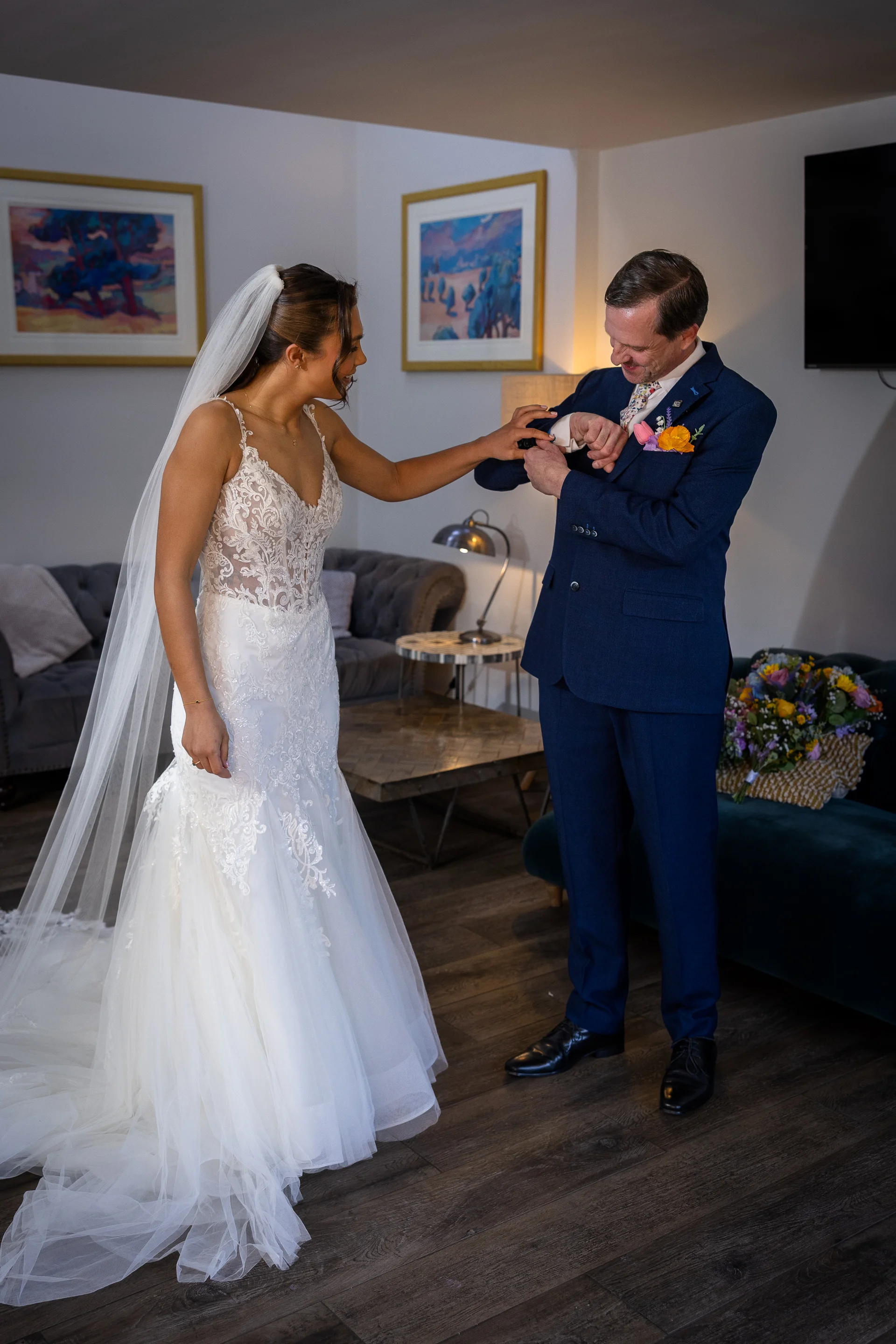 A smiling bride wearing an intricate lace gown and veil gently helps the groom adjust his cufflinks in a light hotel room.