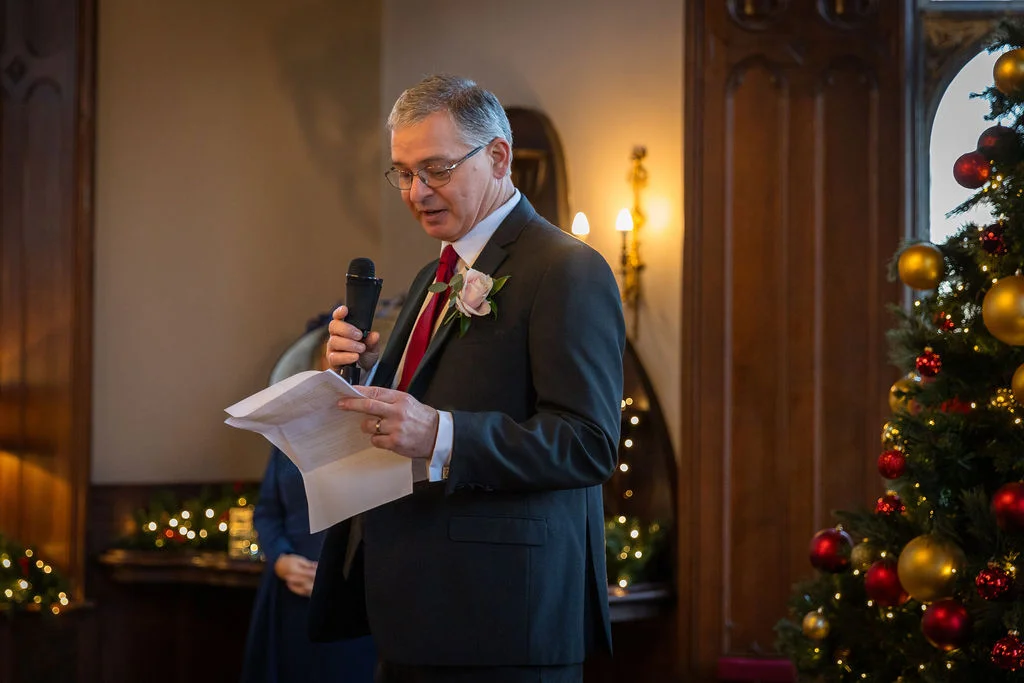 A father of the bride delivers a heartfelt wedding speech holding notes and a microphone beside a decorated Christmas tree.