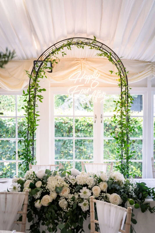 A stunning wedding top table featuring a lush white and green floral runner sits beneath a metal archway wrapped in foliage and a glowing neon sign.