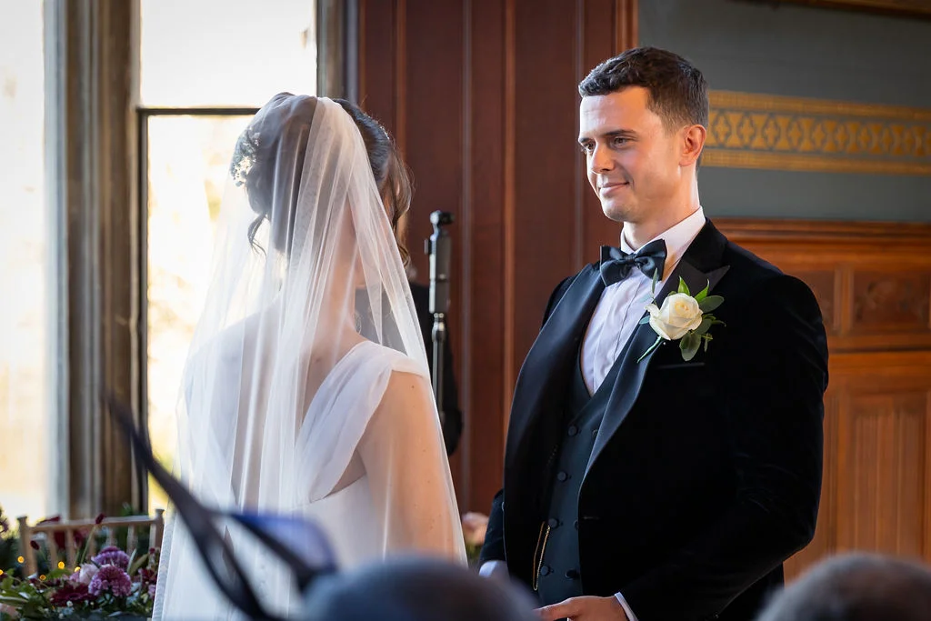 A groom looking lovingly at his bride during their indoor wedding ceremony inside a wood-panelled room.
