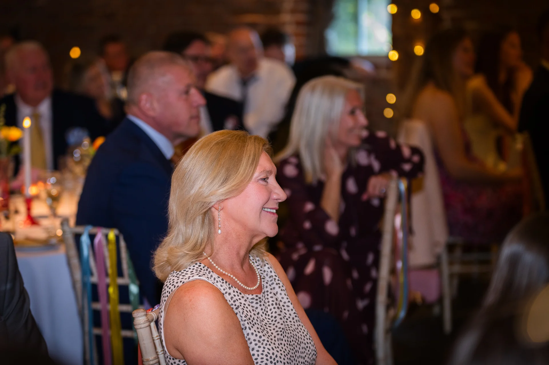 A smiling female wedding guest wearing a polka dot dress and pearl necklace listens intently during the wedding speeches.