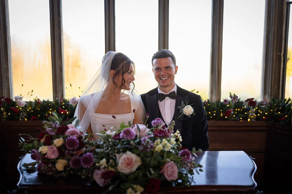 A newlywed couple smiling together behind a large wooden table adorned with rich purple and pink floral arrangements, seated in front of tall panelled windows.