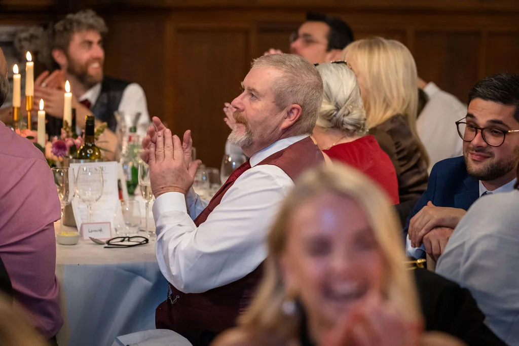 Wedding guests sit around a candlelit round table in a wood-panelled reception room, clapping and laughing during the speeches.