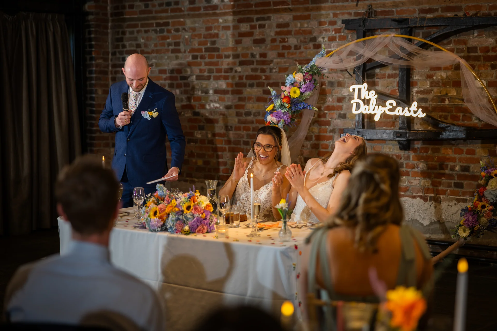 A groom gives a speech at the top table while two brides and wedding guests laugh joyously in an exposed brick industrial venue featuring a glowing neon sign and floral arch.