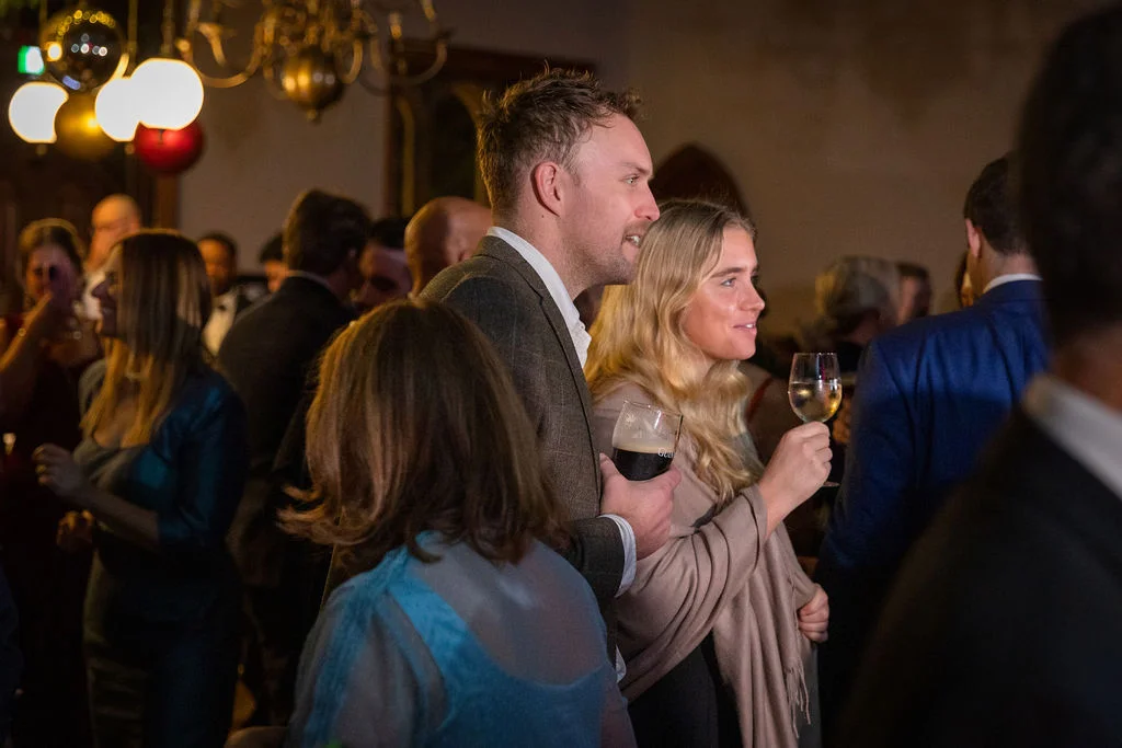 Wedding guests mingle and enjoy drinks during an evening reception in a warmly lit venue hall.