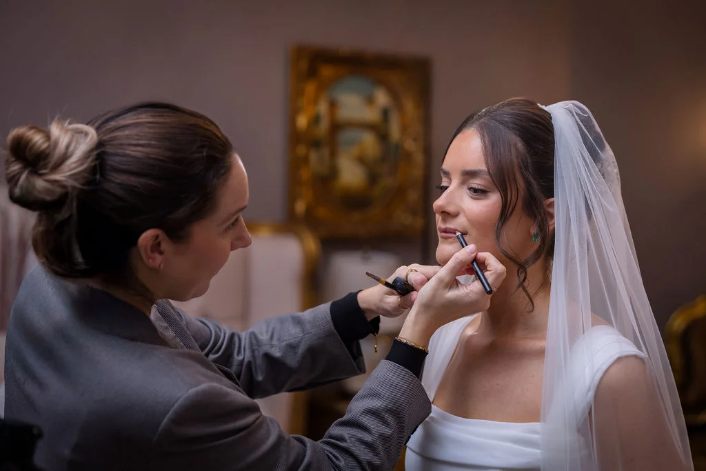 A professional makeup artist carefully applies lip liner to a glowing bride wearing a white gown and veil during morning bridal preparations.
